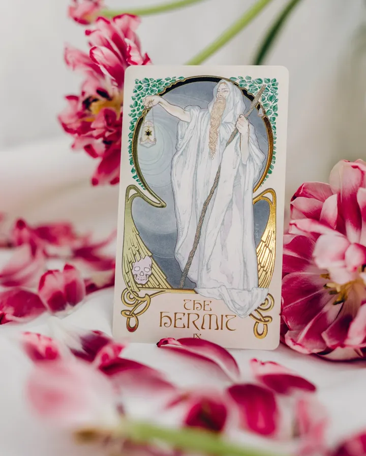 The Hermit tarot card standing upright among pink and white tulips on pale linen