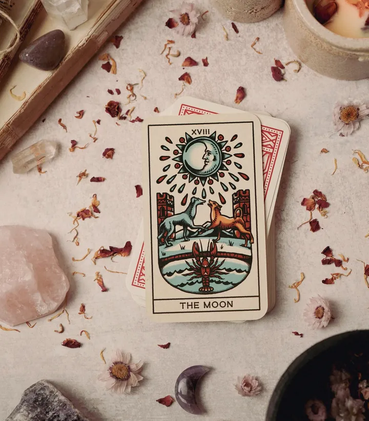 The Moon tarot card on a deck, among rose quartz, moon-shaped stones and dried petals
